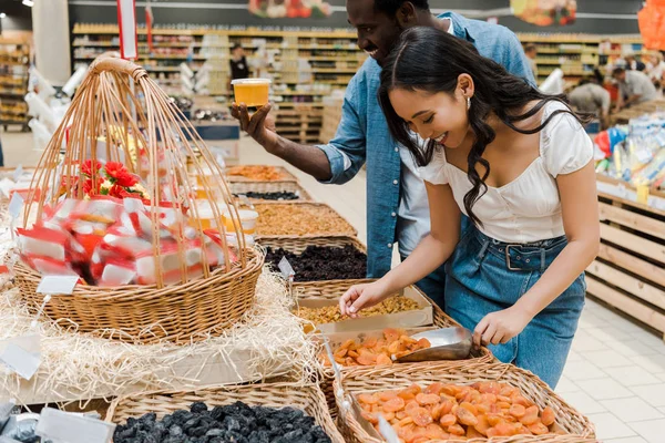 Happy asian woman looking at dried apricots near cheerful african american man holding honey jar in supermarket — Stock Photo