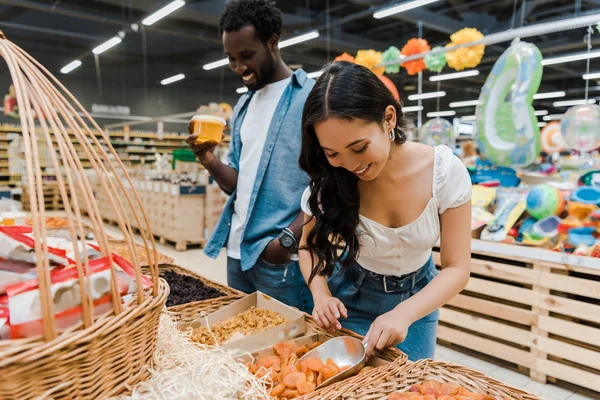 Foco seletivo de mulher asiática feliz olhando para damascos secos perto de homem americano africano alegre segurando frasco de mel no supermercado — Fotografia de Stock