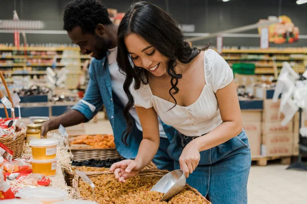 Foco seletivo de mulher asiática feliz olhando para damascos secos perto de homem americano africano alegre no supermercado — Fotografia de Stock