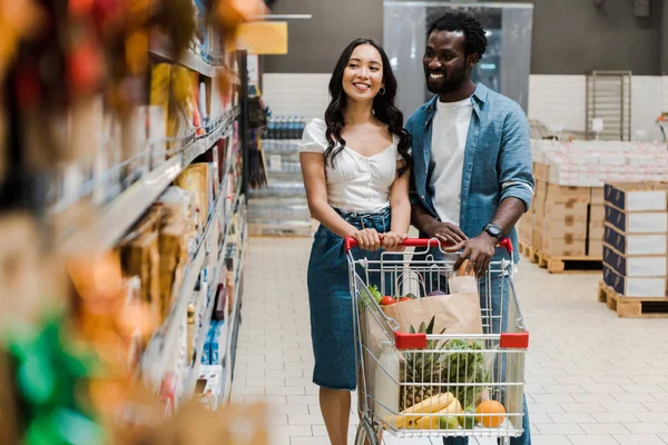 Enfoque selectivo de la mujer asiática feliz de pie cerca alegre afroamericano novio y mirando estantes con comestibles - foto de stock