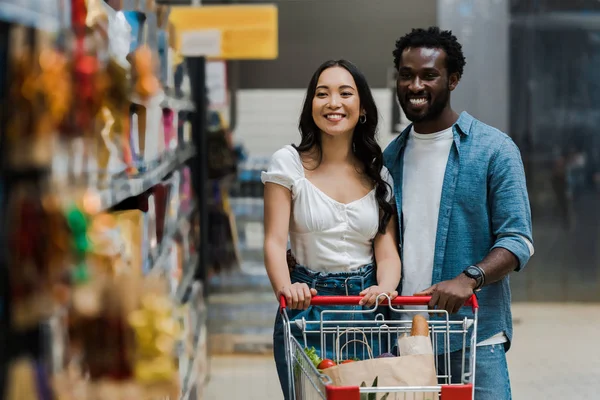 Enfoque selectivo de la mujer asiática feliz y el hombre americano africano alegre mirando estantes en el supermercado - foto de stock