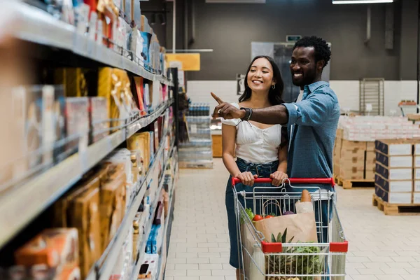 Selective focus of happy asian woman near cheerful african american man pointing with finger at shelves in supermarket — Stock Photo