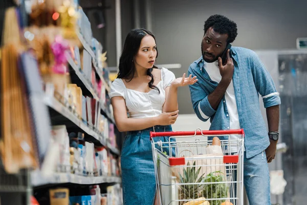 Foyer sélectif de femme asiatique bouleversée gestuelle tout en regardant l'homme afro-américain parler sur smartphone dans un supermarché — Photo de stock