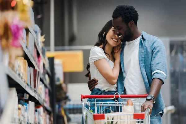 Selective focus of happy african american man hugging asian girl in supermarket — Stock Photo