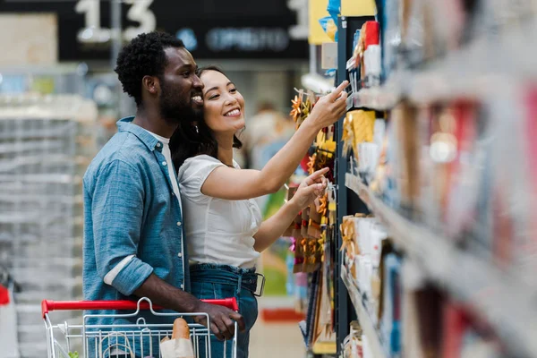Messa a fuoco selettiva di felice uomo africano americano in piedi con donna asiatica che punta con il dito a generi alimentari nel supermercato — Foto stock