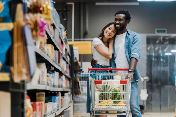 Foco seletivo de homem americano africano alegre abraçando namorada asiática no supermercado — Fotografia de Stock