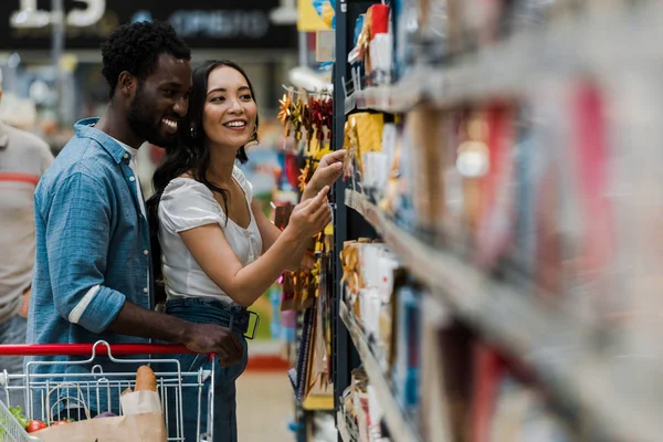 Foco seletivo de homem americano africano alegre em pé com mulher asiática apontando com o dedo para mantimentos no supermercado — Fotografia de Stock