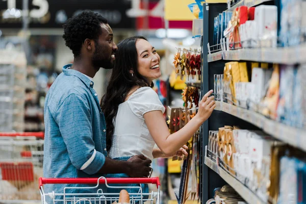 Enfoque selectivo de hombre americano africano alegre de pie con mujer asiática alegre señalando con el dedo a los comestibles en el supermercado - foto de stock