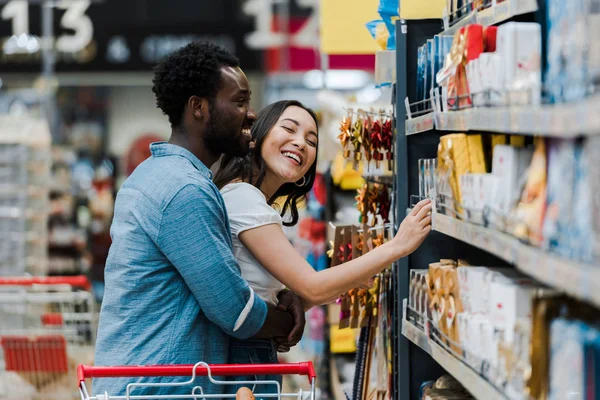 Вибірковий фокус веселого афроамериканця, що стоїть з азіаткою, посміхаючись біля продуктових магазинів у супермаркеті — стокове фото