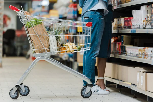 Vista cortada do homem de pé com a mulher perto do carrinho de compras com mantimentos no supermercado — Fotografia de Stock