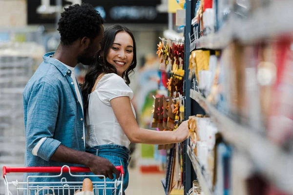 Selektiver Fokus eines fröhlichen afrikanisch-amerikanischen Mannes, der mit einem fröhlichen asiatischen Mädchen in der Nähe von Lebensmitteln im Supermarkt steht — Stockfoto