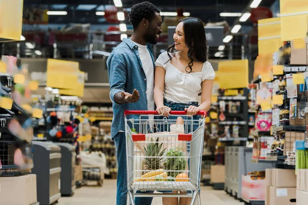 Selective focus of african american man gesturing while looking at asian girl in supermarket — Stock Photo