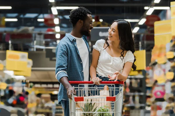 Glücklich afrikanisch-amerikanischer Mann schaut asiatisches Mädchen gestikulierend in der Nähe von Einkaufswagen im Supermarkt an — Stockfoto
