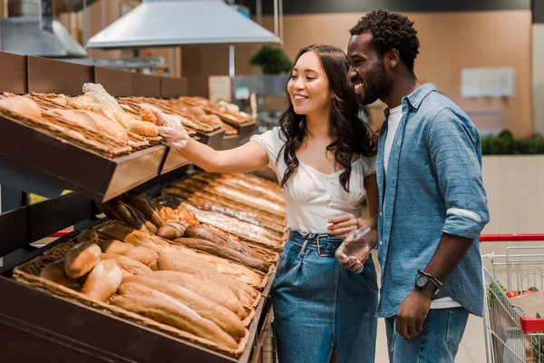 Happy asian woman touching bread while standing near african american man in supermarket — Stock Photo