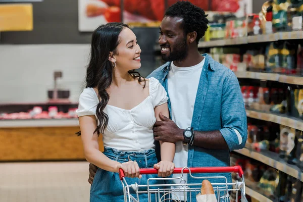 Happy asian woman and handsome african american man looking at each other in supermarket — Stock Photo