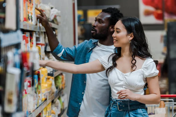 Selektiver Fokus der hübschen Afroamerikanerin, die mit dem Finger in der Nähe attraktiver asiatischer Frauen auf Regale mit Lebensmitteln zeigt — Stockfoto