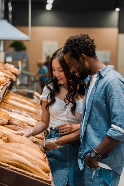 Feliz asiático mujer tocando pan mientras de pie cerca africano americano hombre con mano en bolsillo en supermercado - foto de stock