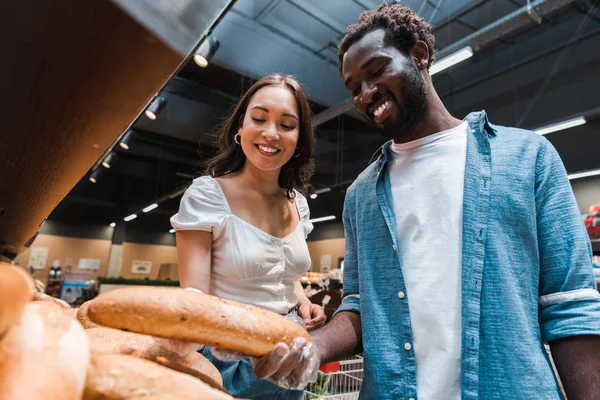 Low-Winkelaufnahme eines afrikanisch-amerikanischen Mannes mit Baguette im Supermarkt in der Nähe einer asiatischen Frau — Stockfoto
