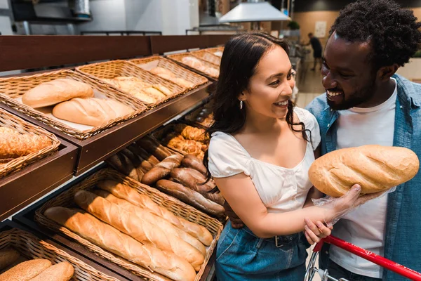 Happy asian woman holding bread near cheerful african american man — Stock Photo