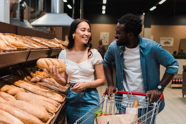 Glücklich asiatische Frau hält Brot in der Nähe fröhlichen afrikanisch-amerikanischen Mann mit Einkaufswagen stehen — Stockfoto