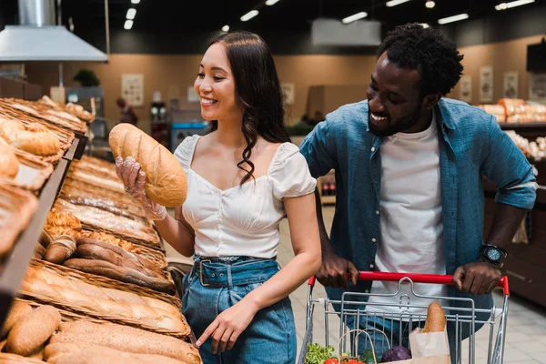 Alegre asiático mulher segurando pão perto africano americano homem de pé com carrinho de compras — Fotografia de Stock