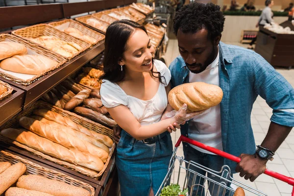 Africano americano hombre mirando pan en mano de alegre asiático mujer - foto de stock