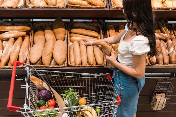 Joven mujer de pie cerca del carrito de la compra mientras sostiene el pan - foto de stock