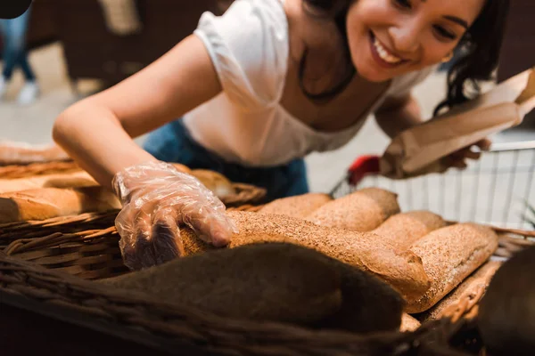 Vista ritagliata di donna felice in guanto vicino al pane nel supermercato — Foto stock