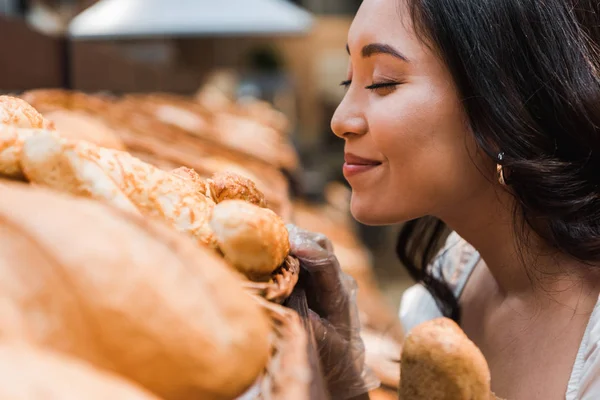 Избирательный фокус веселой азиатской женщины, улыбающейся при запахе хлеба в супермаркете — стоковое фото