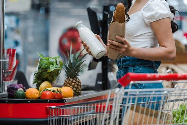 Cropped view of woman holding baguette and glass bottle with milk near supermarket counter — Stock Photo
