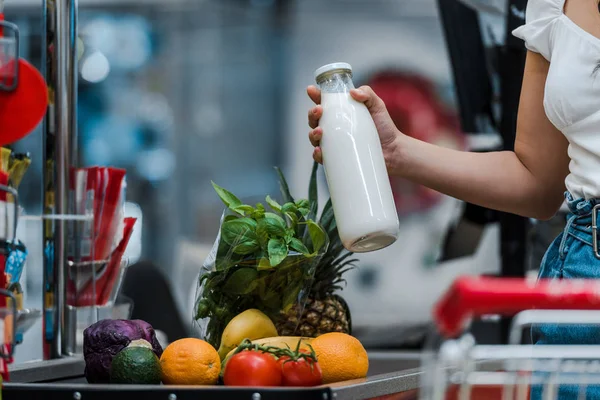 Vista recortada de la mujer sosteniendo botella de vidrio con leche cerca del mostrador del supermercado - foto de stock