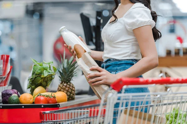 Vista recortada de la joven mujer sosteniendo baguette y botella de vidrio con leche cerca del mostrador del supermercado - foto de stock