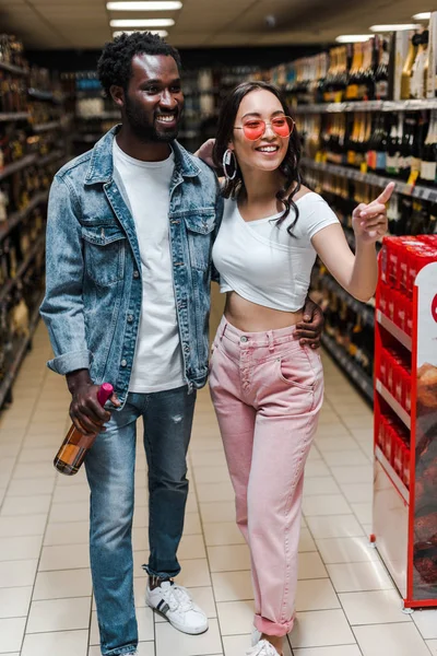 Feliz africano americano hombre celebración botella cerca asiático chica en gafas de sol apuntando con dedo en supermercado - foto de stock