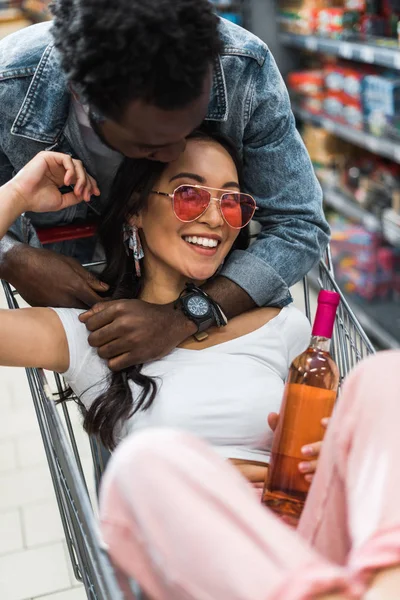 Selective focus of cheerful african american man looking at young asian woman in sunglasses holding bottle and sitting in shopping cart — Stock Photo