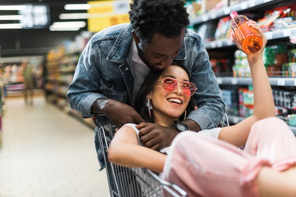 Вибірковий фокус щасливого афроамериканця обіймає азіатську дівчину в сонцезахисних окулярах з пляшкою, що сидить у кошику — стокове фото