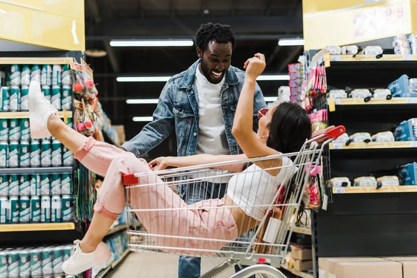 Happy african american man looking at asian girl gesturing while sitting in shopping cart — Stock Photo