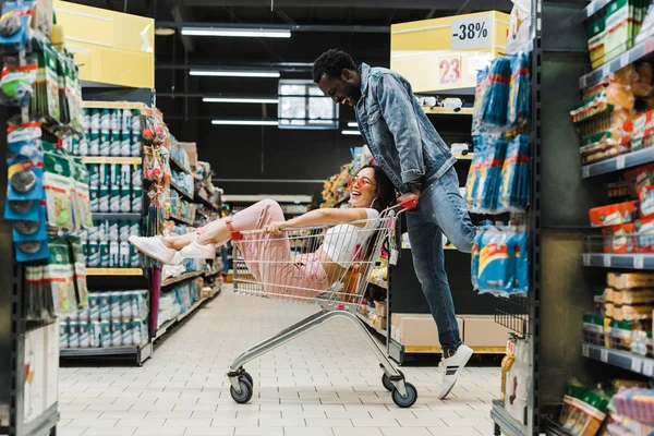 Feliz homem americano africano olhando asiático menina sentado no carrinho de compras no supermercado — Fotografia de Stock