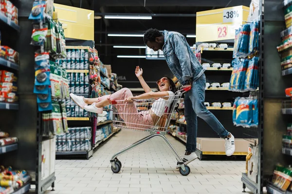 Feliz homem americano africano olhando asiático menina sentado no carrinho de compras e gesticulando no supermercado — Fotografia de Stock
