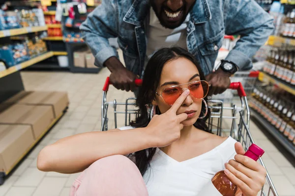 Selettivo fuoco di asiatico ragazza toccare occhiali da sole e holding bottiglia con vino mentre seduta in shopping cart vicino allegro africano americano uomo — Foto stock