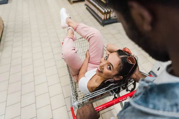 Вид зверху на афроамериканця біля веселої азіатської дівчини, що сидить у кошику в супермаркеті — стокове фото