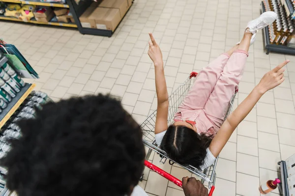 Vue aérienne d'un homme afro-américain près d'une fille assise dans un panier et faisant un geste dans un supermarché — Photo de stock