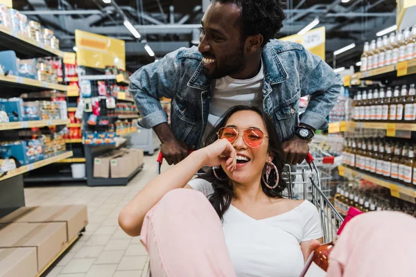 Вибірковий фокус щасливої азіатської дівчини сонцезахисні окуляри і тримає пляшку з вином, сидячи в кошику біля веселого афроамериканця в магазині — стокове фото