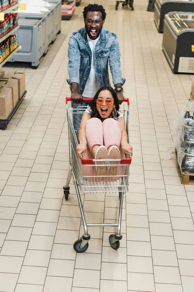 Hombre afroamericano feliz caminando con chica asiática en gafas de sol sentado en el carrito de compras - foto de stock