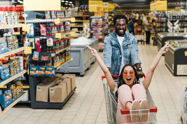 Excited african american man near happy asian girl in sunglasses gesturing while sitting in shopping cart — Stock Photo