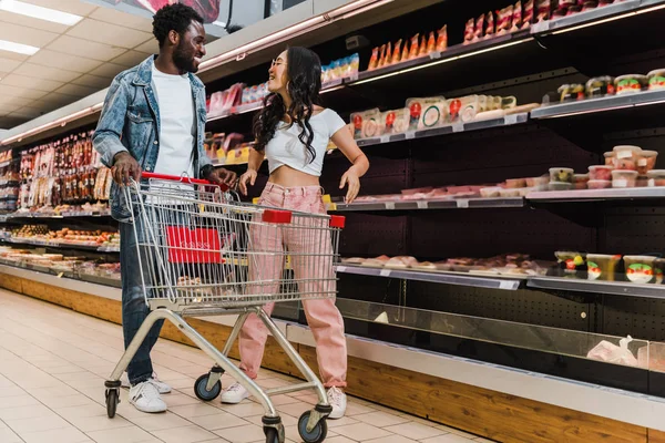 Sorridente afro-americano homem de pé perto feliz asiático menina em óculos de sol e carrinho de compras — Fotografia de Stock