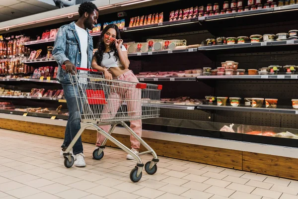 Felice uomo africano americano in piedi vicino elegante ragazza asiatica toccare occhiali da sole nel supermercato — Foto stock