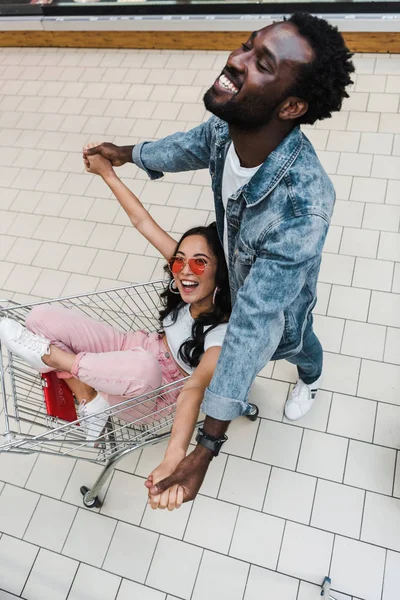 Vista aérea de la chica asiática feliz en gafas de sol sentado en el carrito de la compra y cogido de la mano con el hombre afroamericano guapo - foto de stock