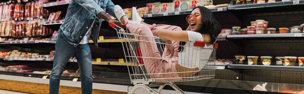 Panoramic shot of happy asian girl in sunglasses sitting in shopping cart near african american man — Stock Photo