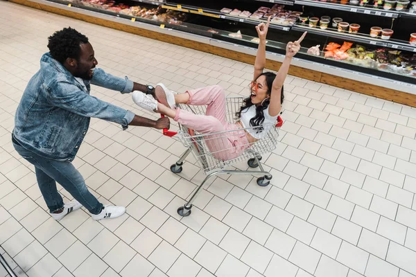 Opinião aérea a menina asiática feliz nos óculos de sol que gesticula ao sentar-se no carro de compra e em olhar o homem do americano africano — Fotografia de Stock