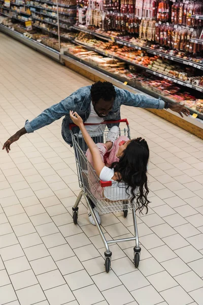 Menina asiática em óculos de sol sentado no carrinho de compras perto de homem americano africano com as mãos estendidas — Fotografia de Stock
